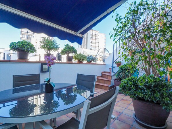 A glass-top table with chairs, surrounded by potted plants, under a blue awning. - Benito Apartment by Hello Homes Sitges (Sitges)