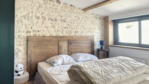 A bedroom with a rustic stone wall, wooden beams, a bed with white bedding, and a small bedside table with a lamp.