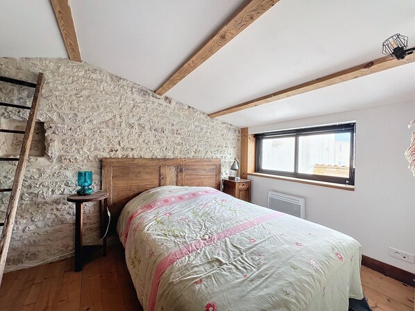 Une chambre avec un mur en pierre, des poutres en bois, un lit avec une literie à motifs floraux, une fenêtre donnant sur un beau paysage et une table de chevet avec une lampe.
