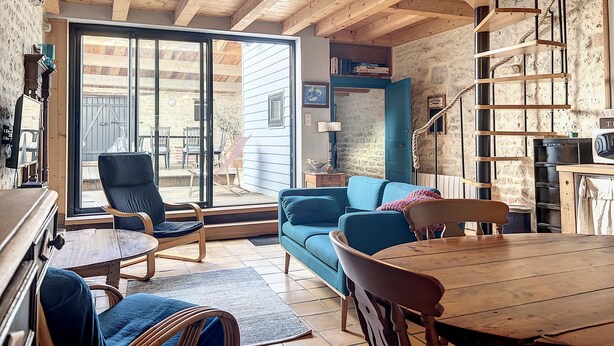 A cozy living room with a wooden ceiling, a blue sofa, a wooden dining table, and a staircase with a rope railing.