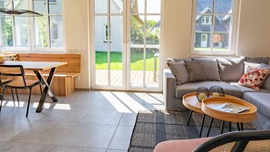 Una sala moderna con un sofá, una mesa de comedor y un banco. Hay ventanas amplias y una puerta corrediza de vidrio que da a un patio.