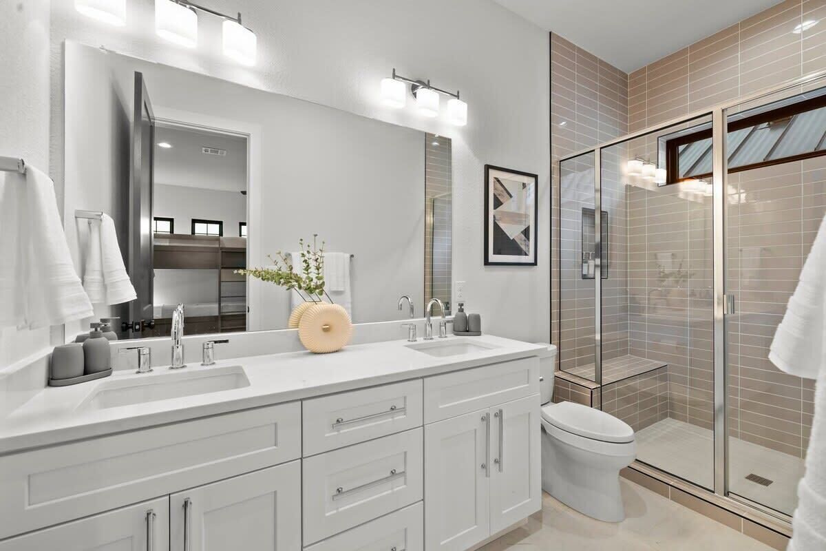 A modern bathroom with a large mirror, two sinks, a toilet, and a glass-enclosed shower.