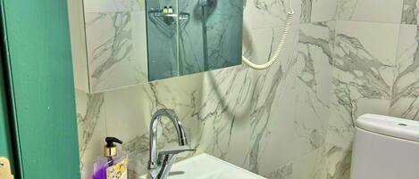 A bathroom with a marble-tiled wall, a white sink with a soap dispenser, a toilet, and a glass-enclosed shower area.