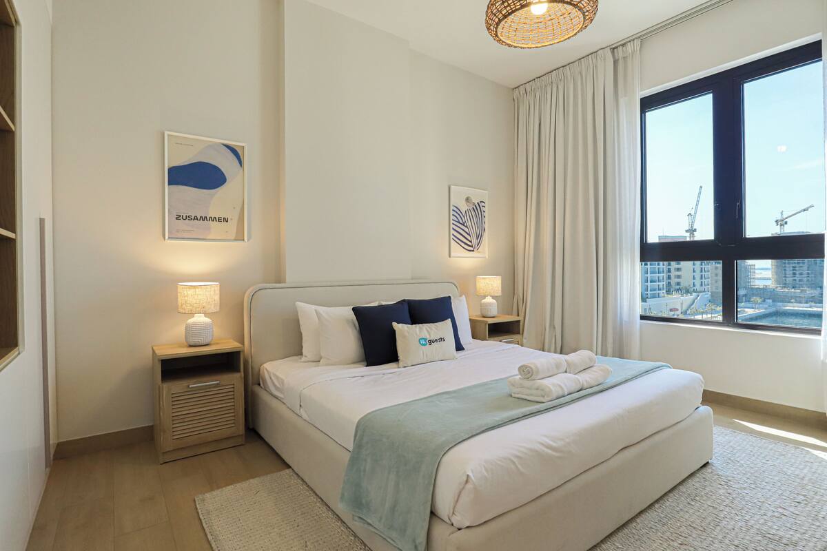 A modern hotel room with a large bed, two bedside tables with lamps, a view of the city through a large window, and artwork on the walls.