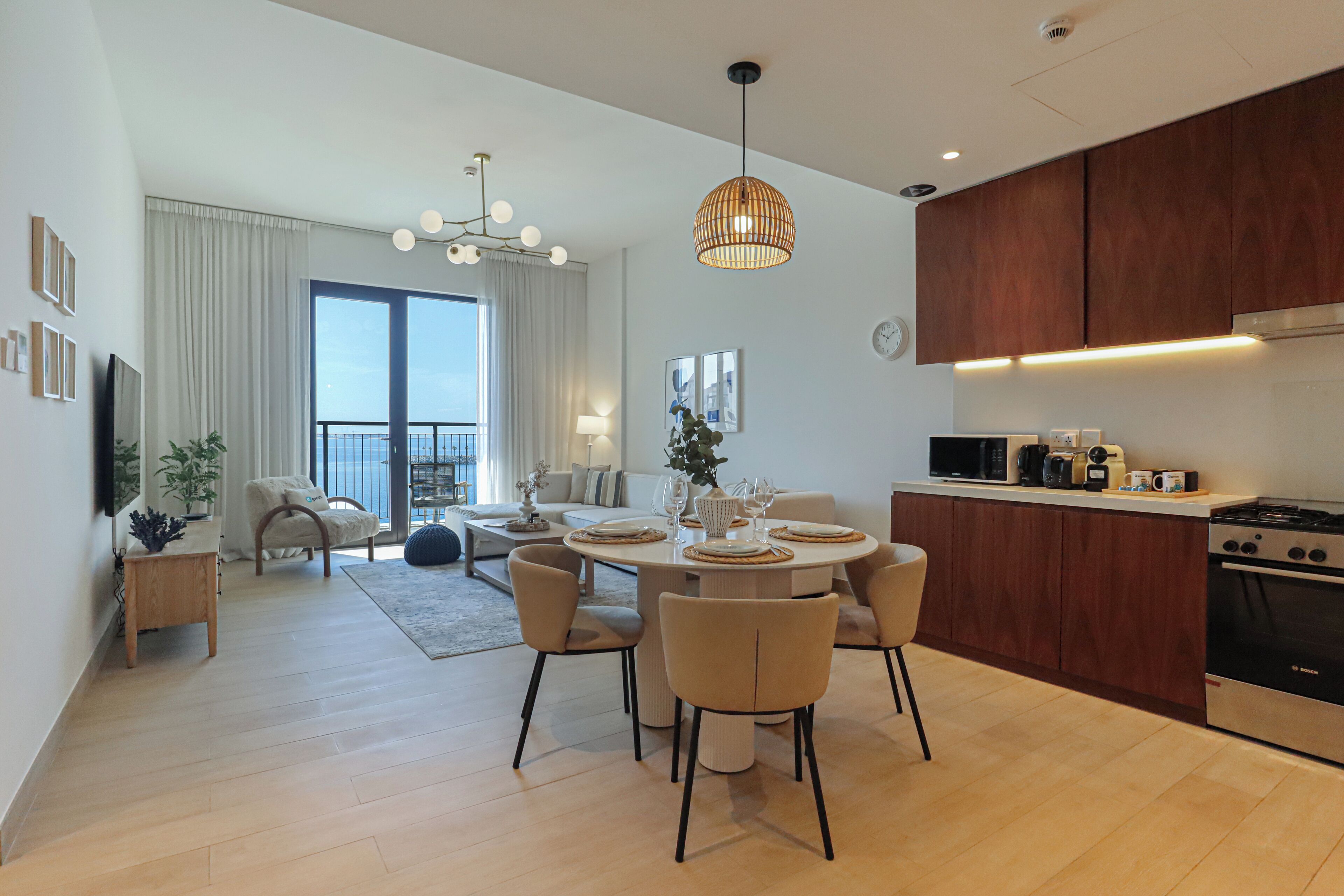 A modern kitchen with wooden cabinets, a dining area with a round table and chairs, and a large window with curtains.