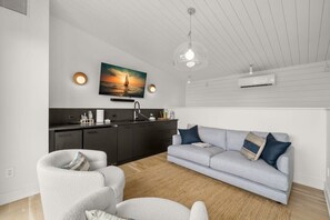 A modern living room with a grey sofa, white armchairs, a kitchen area with dark cabinets, and a large painting of a sunset over water on the wall.
