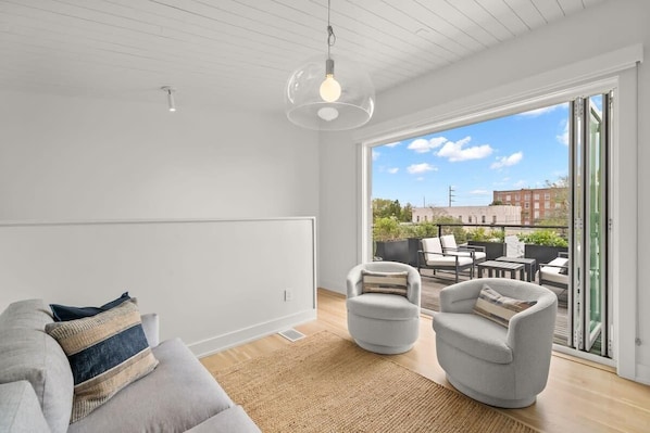 Ein modernes Wohnzimmer mit Sofa, Sesseln, einem großen Fenster und einem Balkon mit Gartenmöbeln.