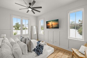 A bedroom with a bed, a ceiling fan, a painting of a sunset, and large windows.