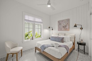 Une chambre à coucher comprenant un lit, une chaise, une fenêtre avec des stores, un ventilateur de plafond et une œuvre d’art accrochée au mur.