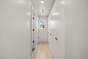 A narrow hallway with white walls and wooden flooring, leading to a door with a window at the end.