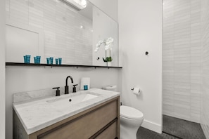 Une salle de bain moderne avec un lavabo blanc, un robinet noir et un miroir au-dessus. On trouve des toilettes sur la droite et un espace douche avec un sol carrelé.