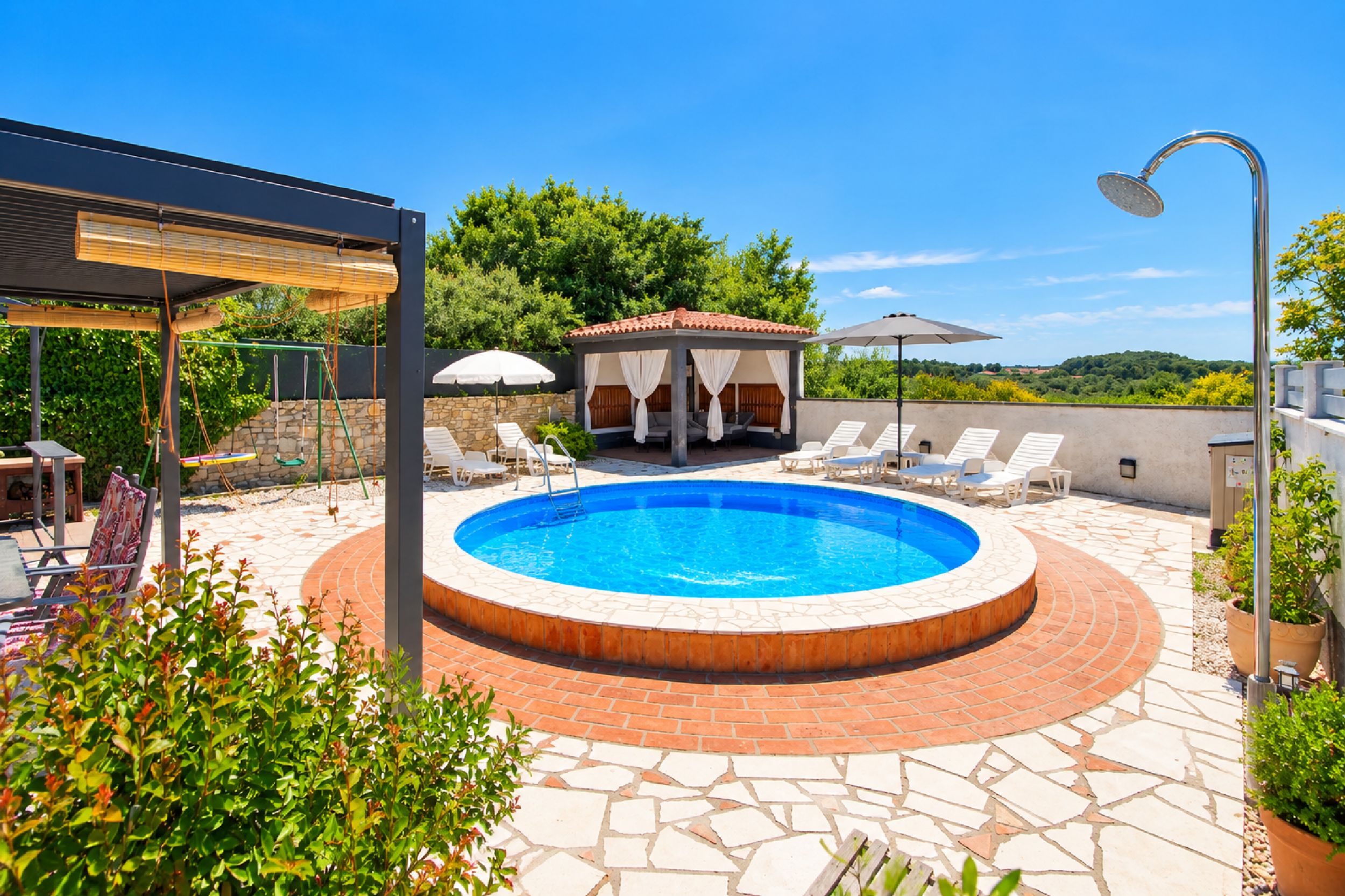 A circular swimming pool with a brick edge, surrounded by lounge chairs and a covered seating area.