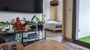 Un salon moderne avec une table basse en bois, un téléviseur fixé au mur, un lit avec un matelas blanc et une tête de lit en bois, ainsi que plusieurs plantes en pot.