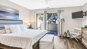 A bedroom with a large bed, a TV mounted on the wall, a desk with a chair, and a balcony with a view of the outdoors.