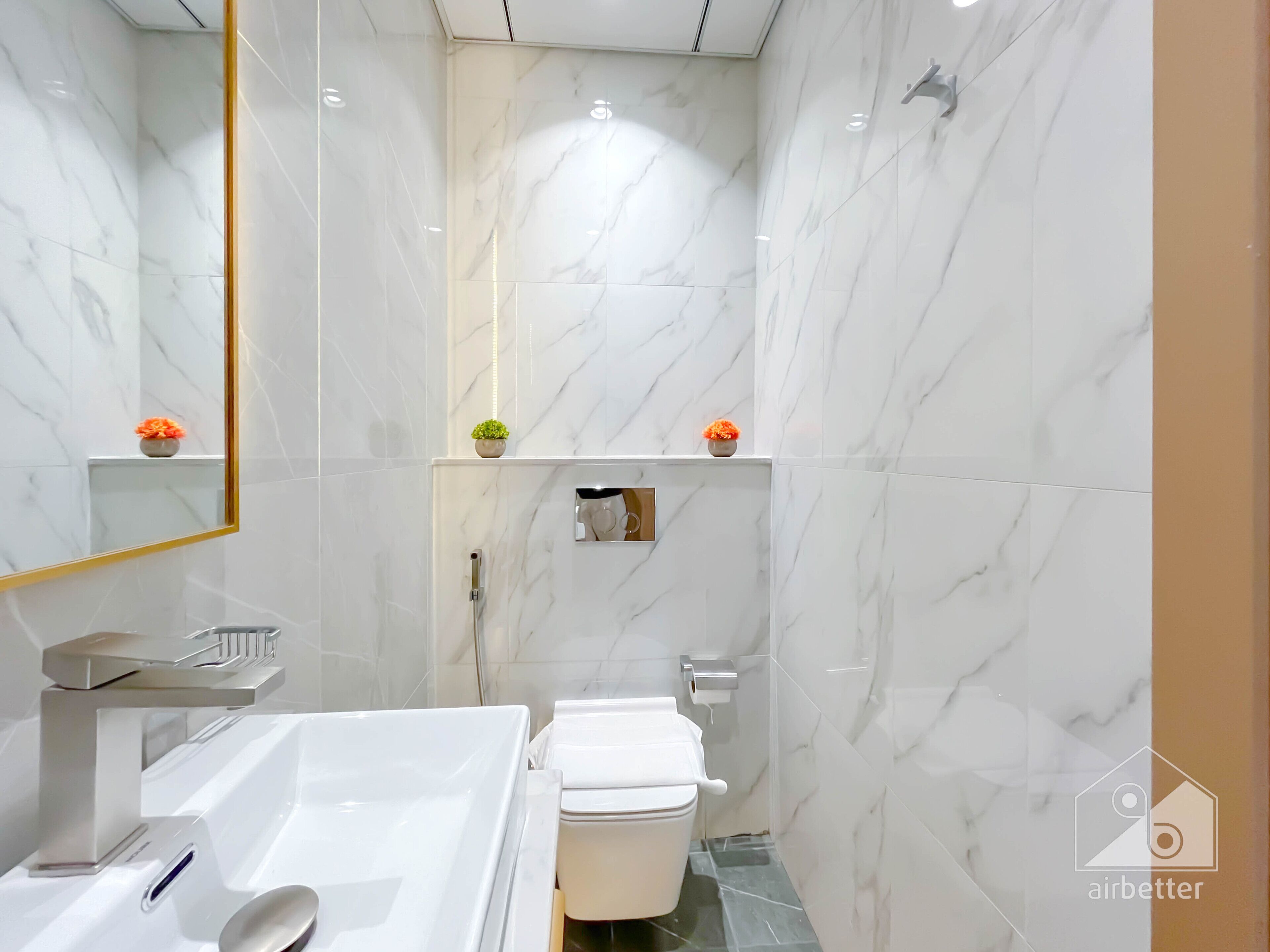 Une salle de bain moderne avec un mur en marbre blanc, un grand miroir, une baignoire blanche, des toilettes et une petite étagère sur laquelle est posée une plante.