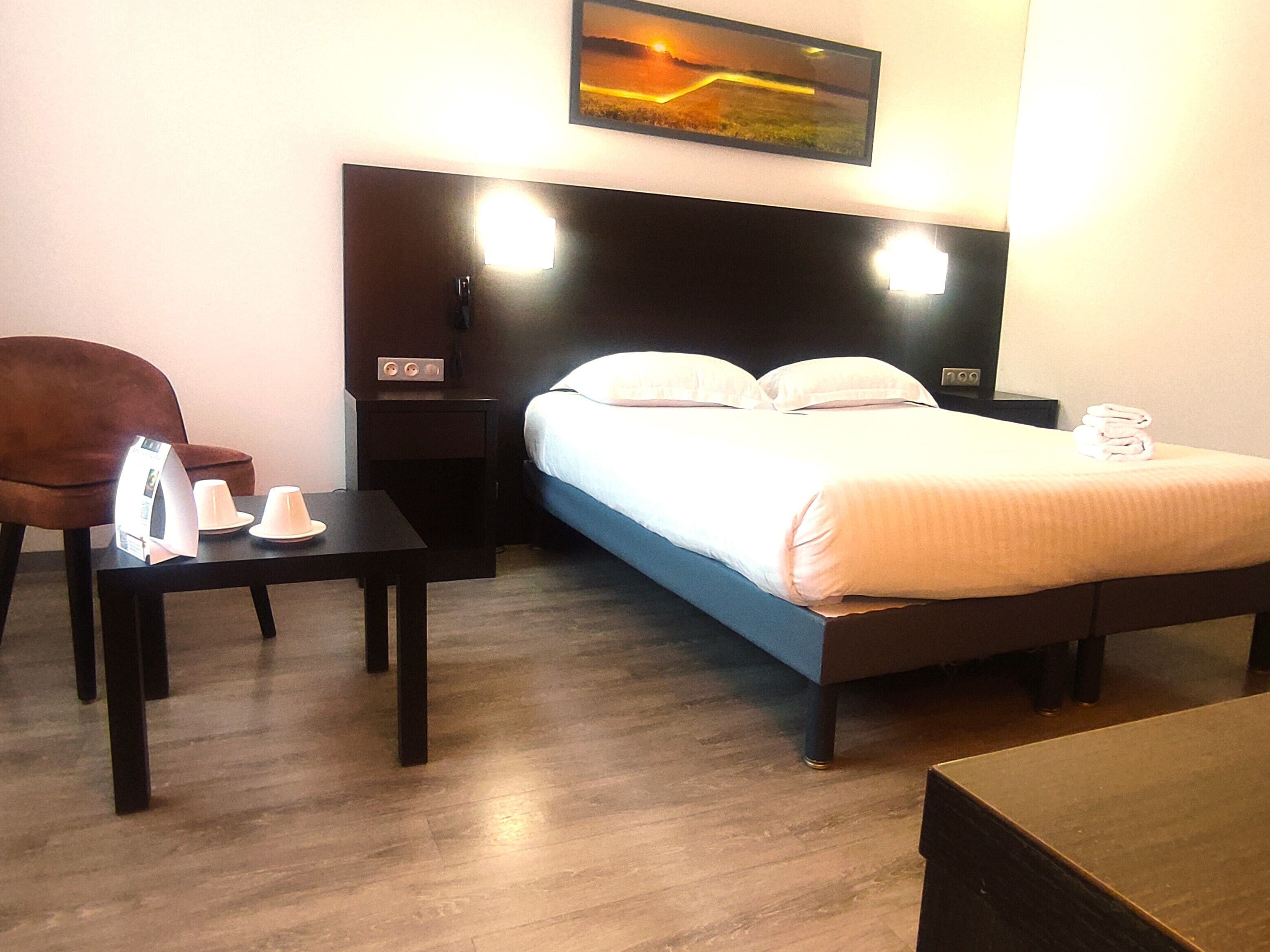 A hotel room with a bed, bedside table, chair, and a painting on the wall.