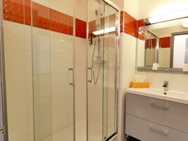 Un baño moderno con ducha de vidrio, paredes con azulejos blancos y rojos, un lavamanos blanco con espejo y una caja de pañuelos de madera.