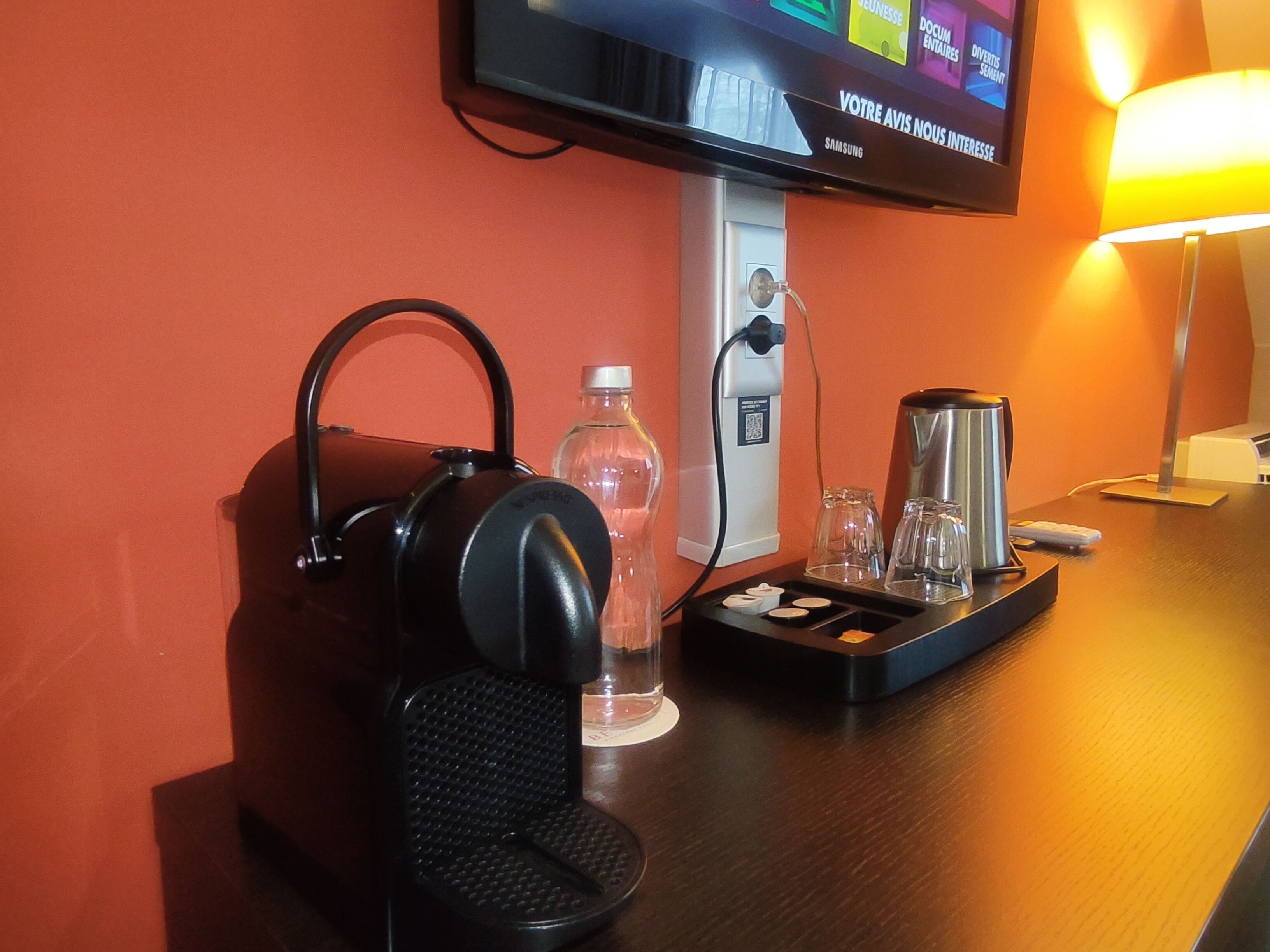 Hotelkamer met een televisie waarop een menu staat, een koffiezetapparaat, een waterfles en een koptelefoon op een houten tafel.