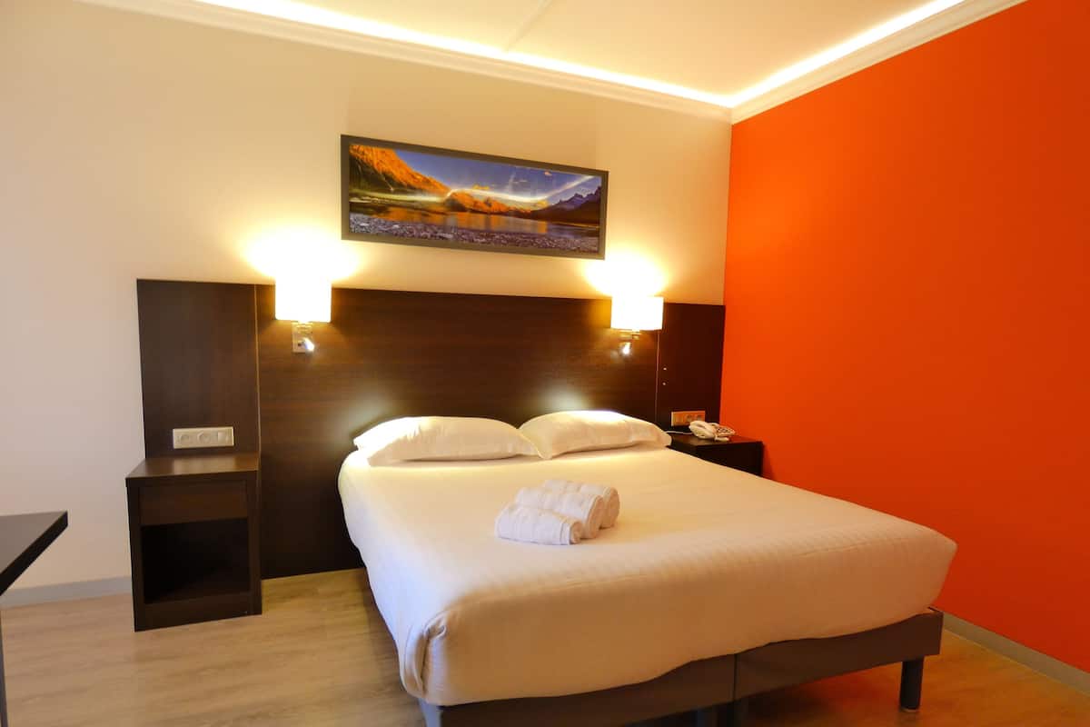 A hotel room with a large bed, two bedside lamps, a nightstand with a phone, a painting on the wall, and a wooden floor.