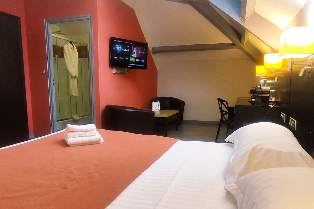 A hotel room with a bed, a desk, a chair, a TV mounted on the wall, and a bathroom visible through an open door.