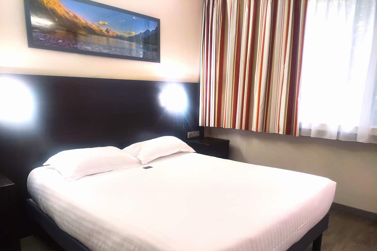 A neatly made bed with white bedding, a dark headboard, and a painting of a mountain landscape above it.