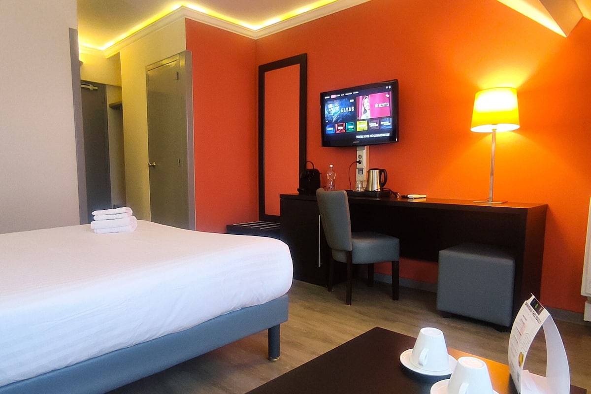 A hotel room with a bed, a desk, a chair, a TV, a lamp, and a coffee table with cups and saucers.