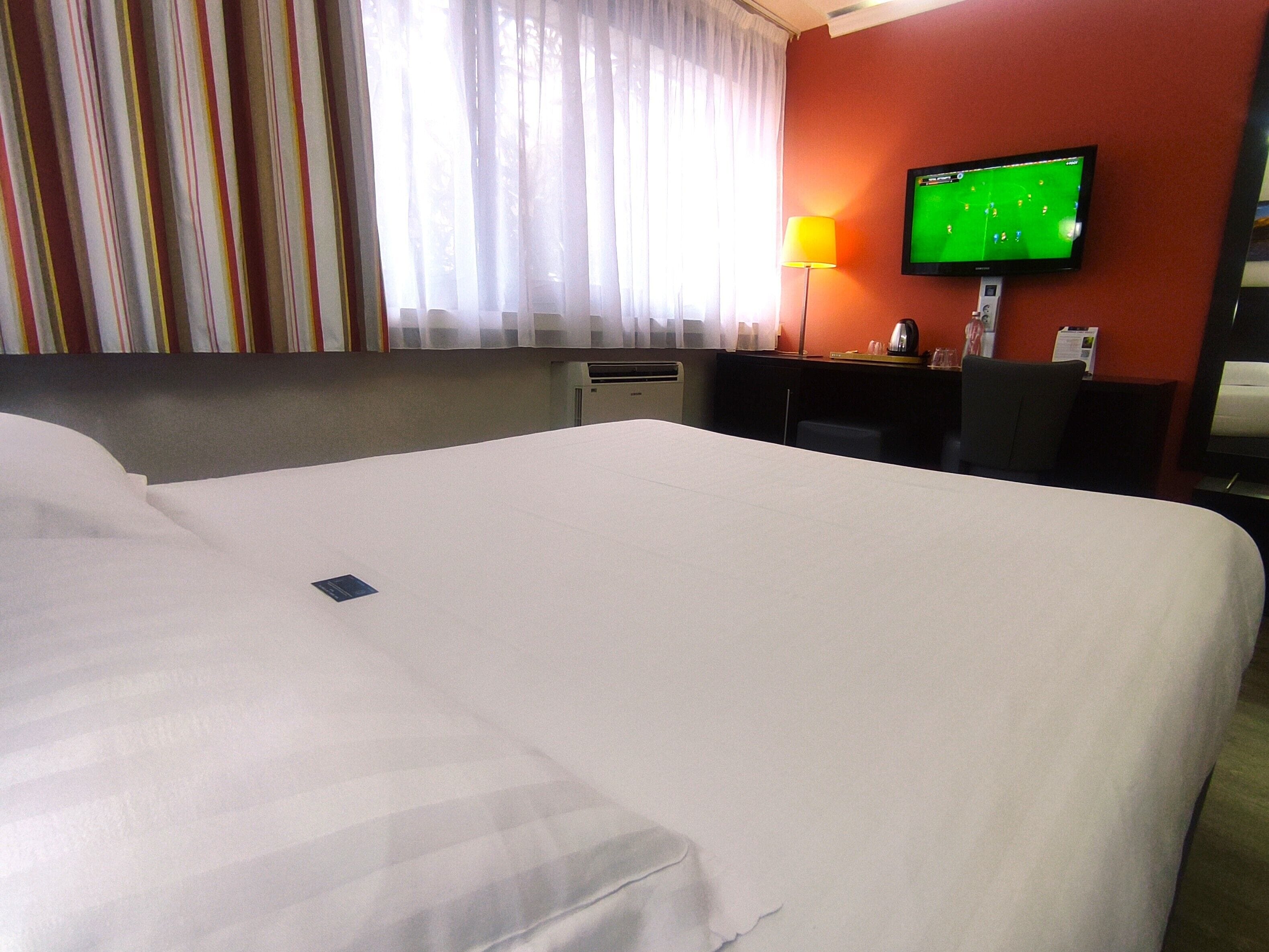 Una habitación de hotel con una cama grande, un escritorio con una computadora, un televisor y una ventana con cortinas.