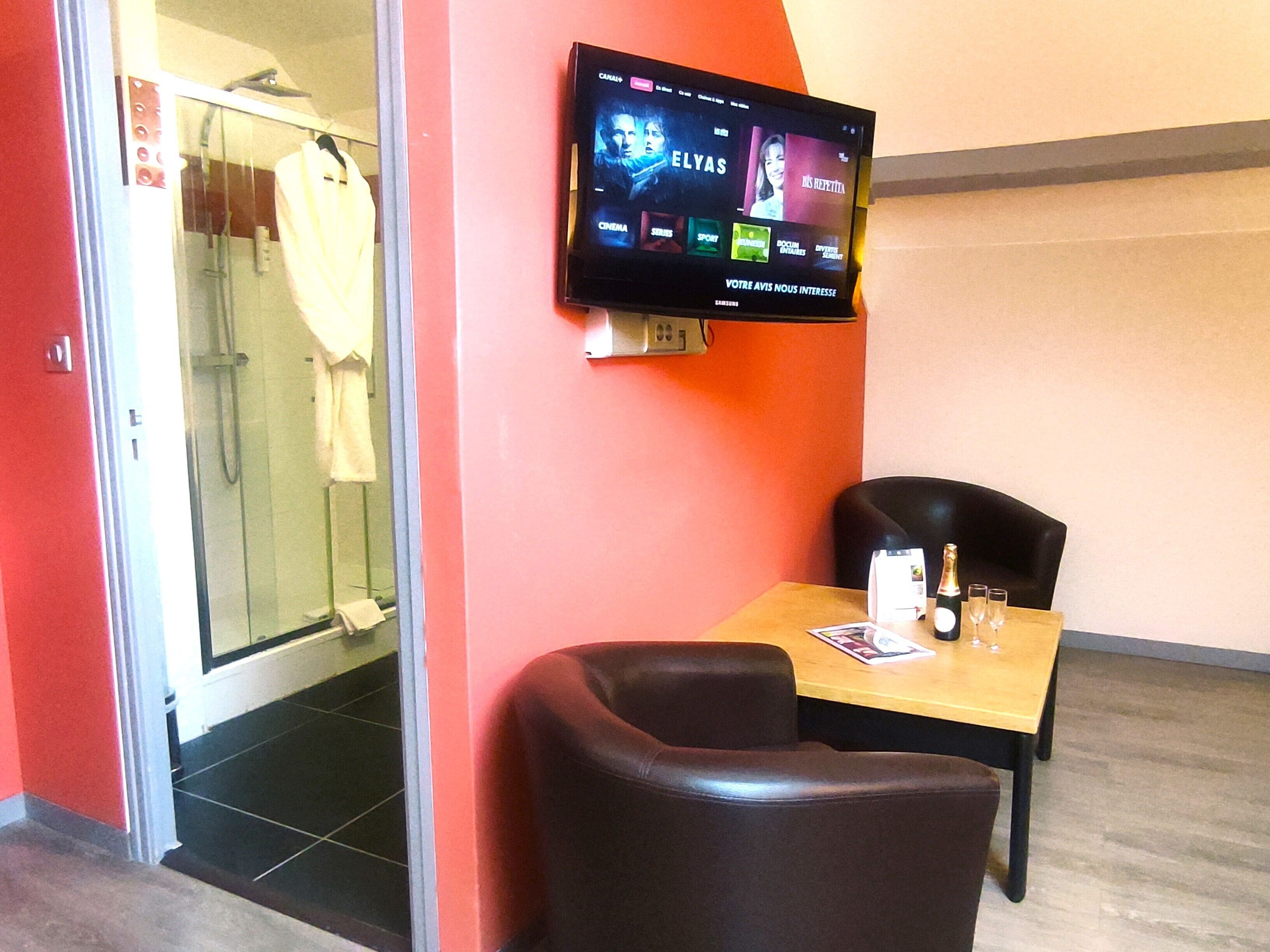 Ein Hotelzimmer mit an der Wand befestigtem Fernseher, der eine Auswahl an Filmen zeigt. Im Duschbereich befindet sich eine Glastür und ein Handtuch hängt daran. Ein Tisch mit zwei Stühlen steht in der Nähe der Wand, und darauf befinden sich eine Flasche und Gläser.