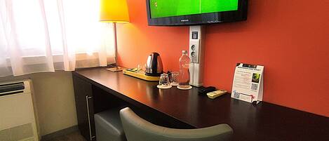 A hotel room with a TV mounted on the wall displaying a soccer game, a desk with a lamp, a coffee maker, and a remote control.
