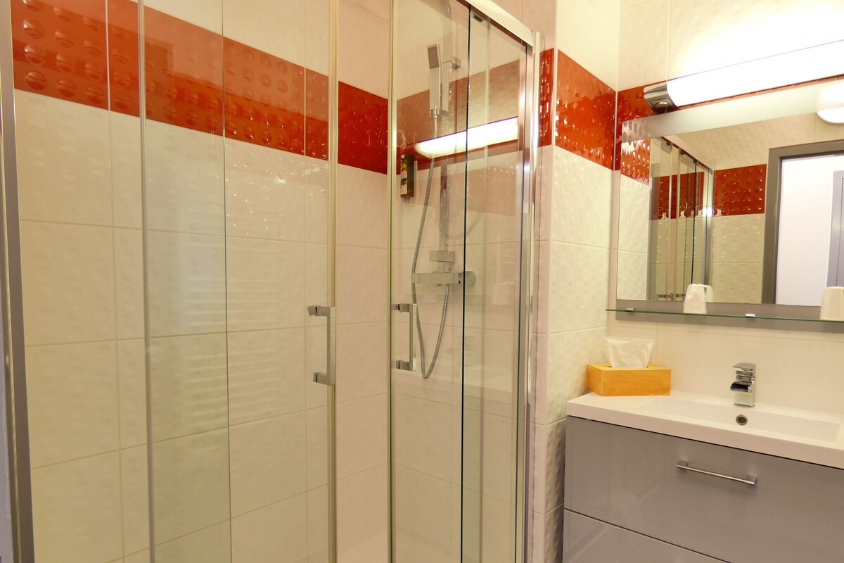 Un baño moderno con ducha de vidrio, paredes con azulejos blancos y rojos, un lavamanos blanco con espejo y una caja de pañuelos de madera.