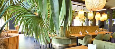 Interior de restaurante com assentos estofados em verde, mesas de madeira e luminárias de vime suspensas.