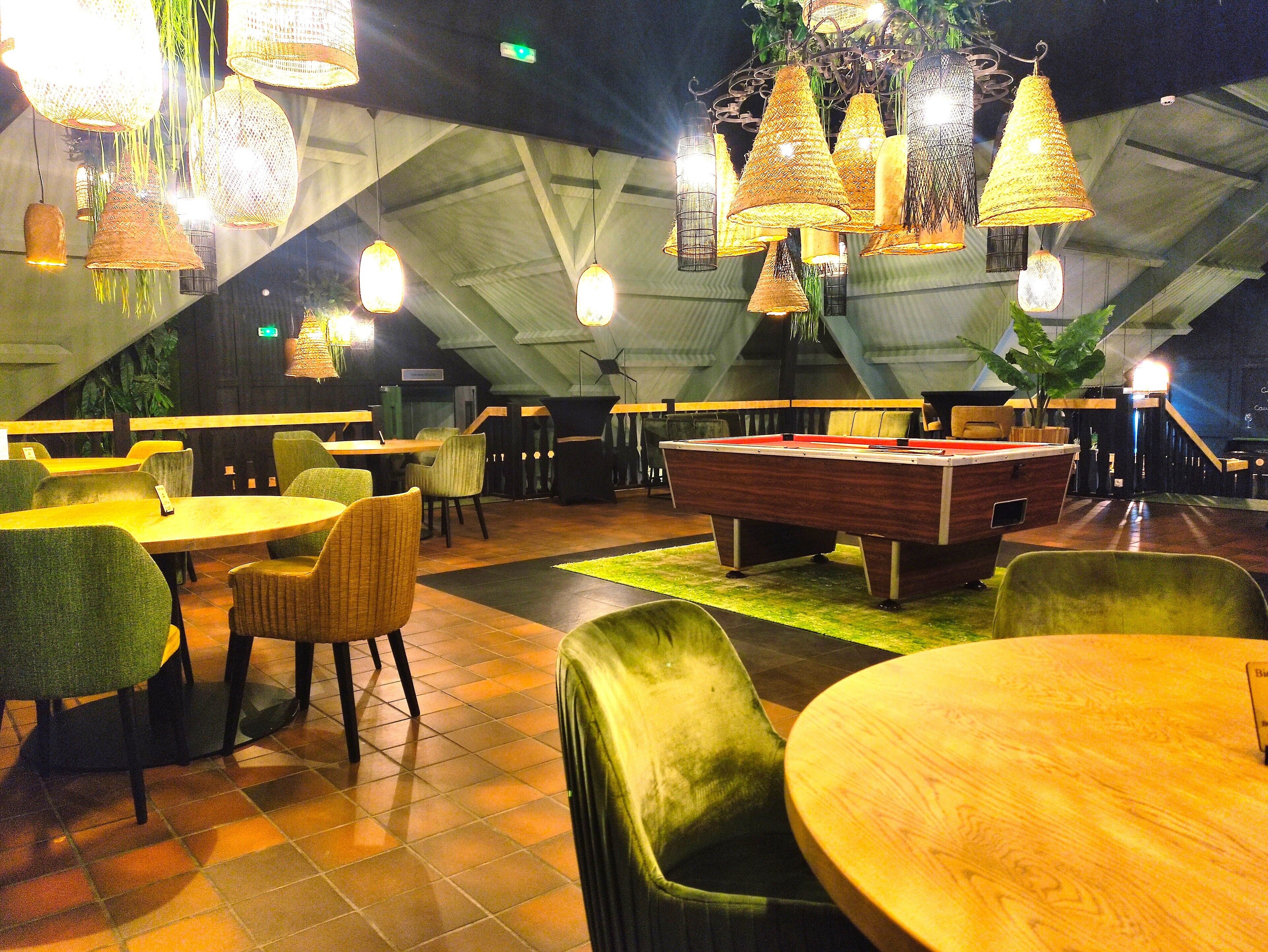 Un espacio interior acogedor con mesa de billar, sillas tapizadas en verde y lámparas colgantes.