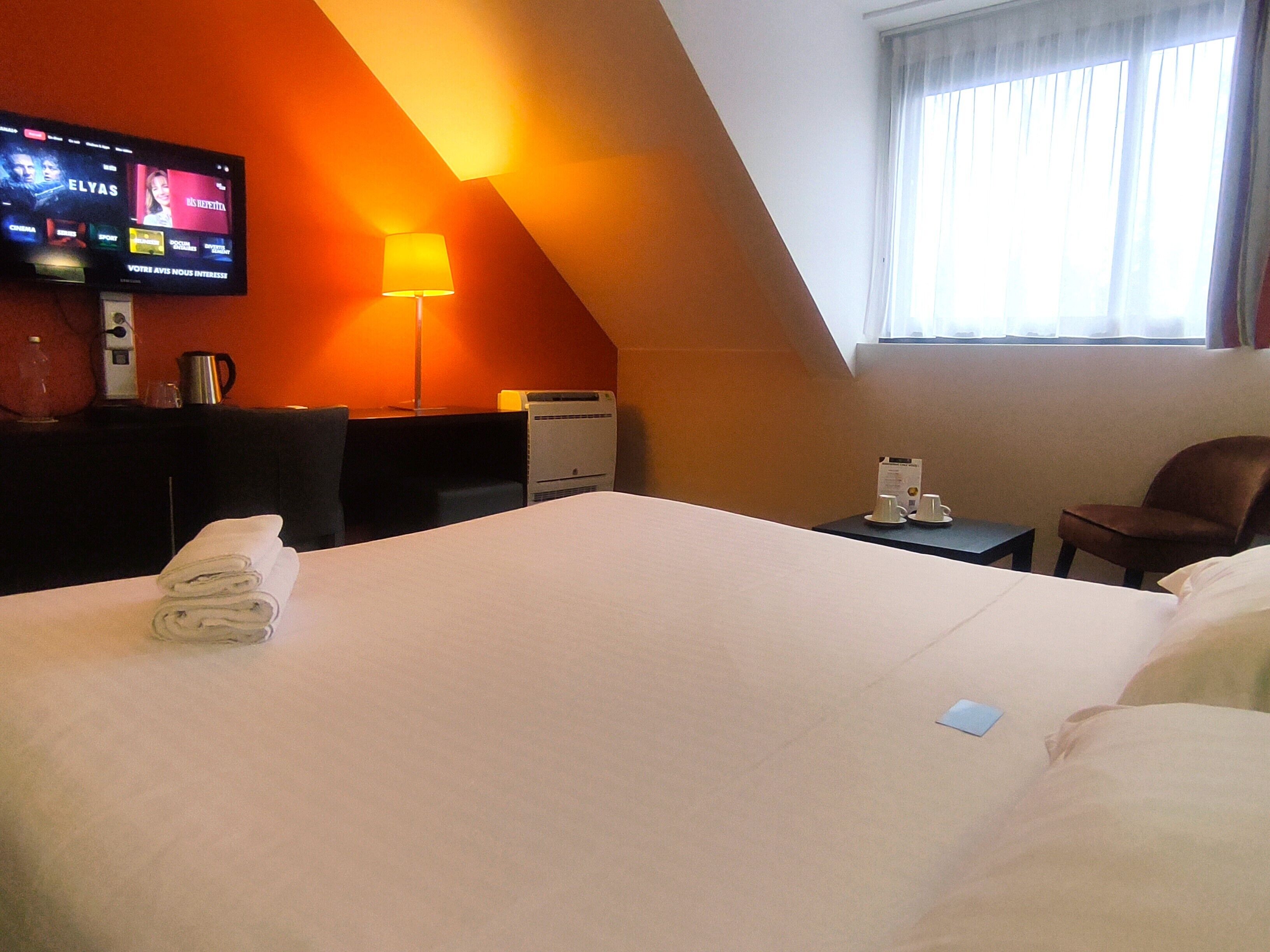 A hotel room with a large bed, a desk with a lamp, a television on the wall, and a window with curtains.