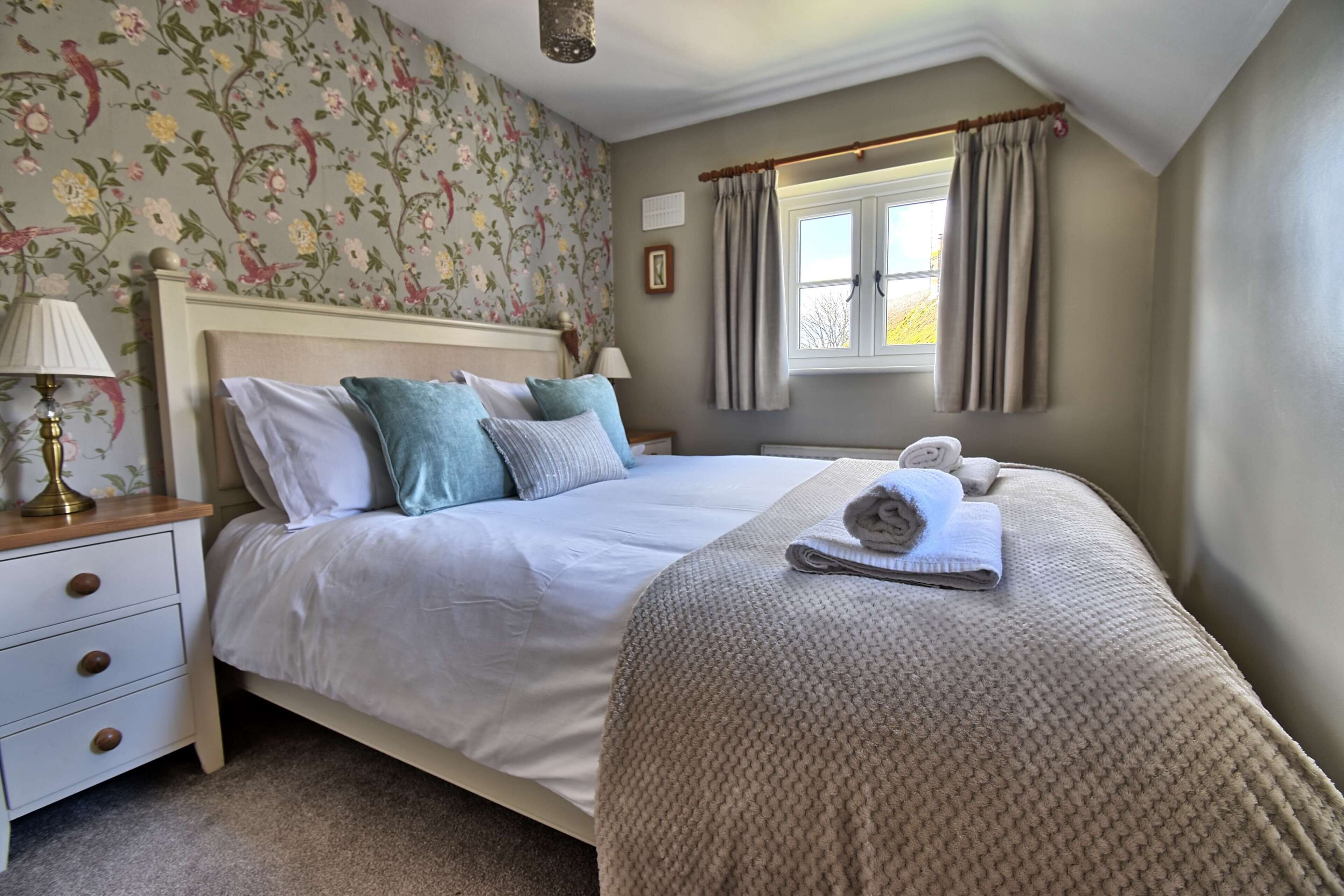 A bedroom with a bed, nightstand, lamp, window with curtains, and a floral wallpaper.