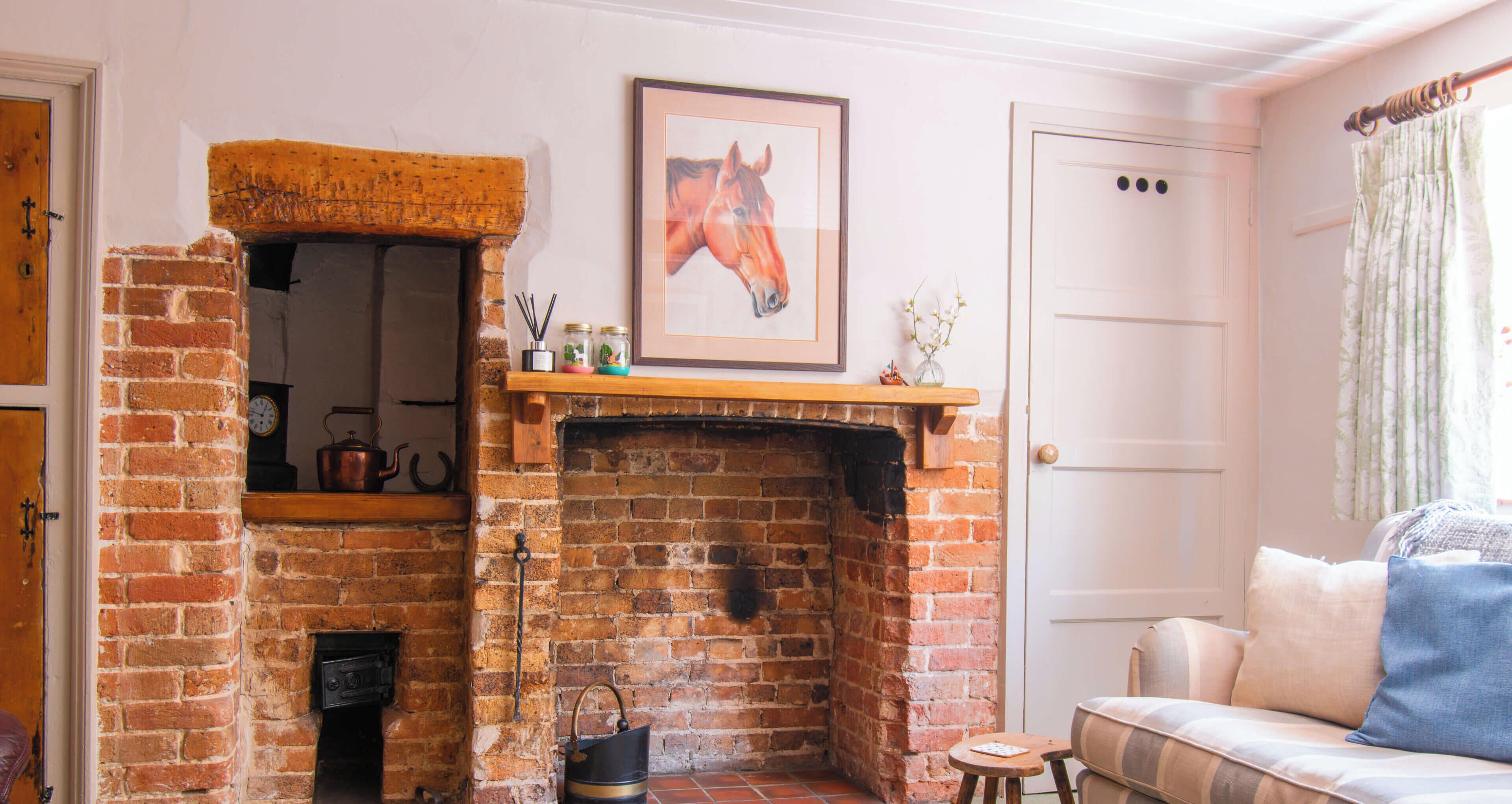 A room with a brick fireplace, a framed horse painting, a door, a window with curtains, and a sofa.
