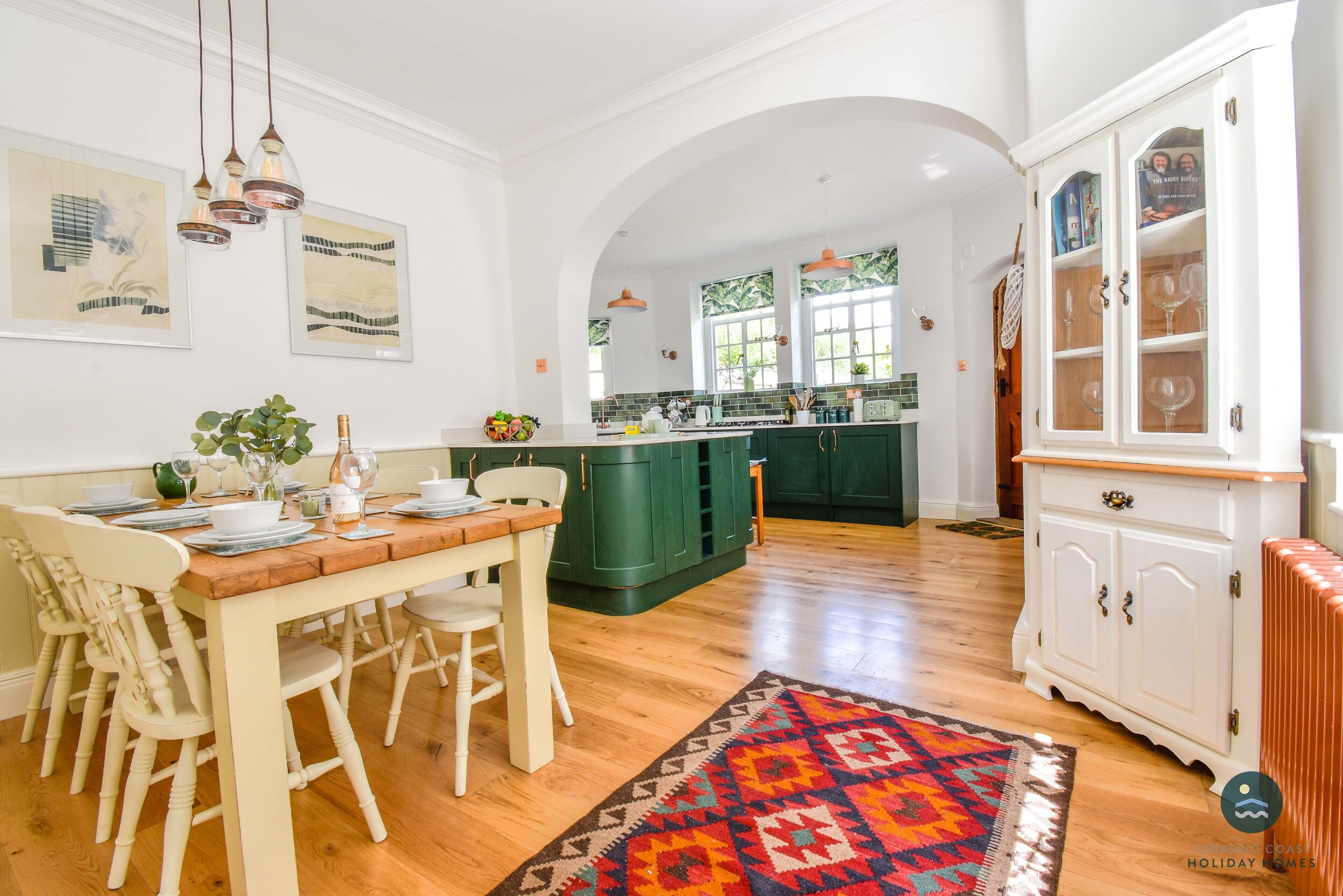 A spacious kitchen with a central island, wooden dining table with chairs, and a patterned rug.