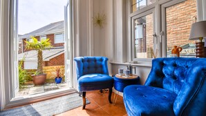 Une pièce avec un canapé en velours bleu, un fauteuil assorti et une petite table d’appoint. Une fenêtre donne sur un bâtiment en briques et sur un palmier à l’extérieur.