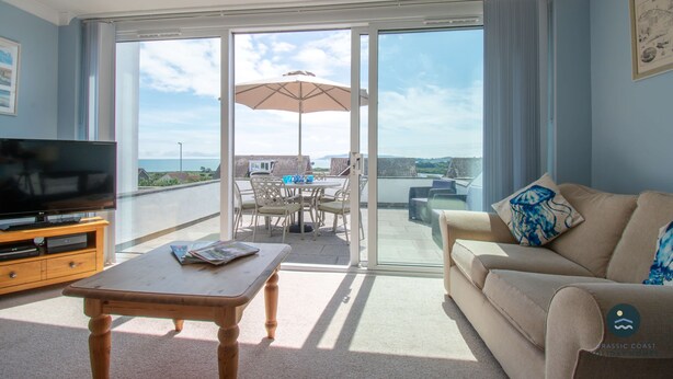 A living room with a TV, a wooden table, a beige sofa, and a balcony with a table and chairs. - Pebble House - Sea views, parking, family friendly (Weymouth)