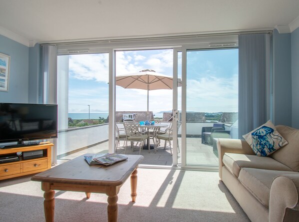 A living room with a TV, a wooden table, a beige sofa, and a balcony with a table and chairs.