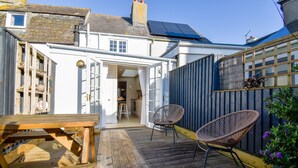 Une terrasse en bois avec une table et deux chaises en osier, qui mène à un bâtiment blanc doté d’un toit bleu et de panneaux solaires.
