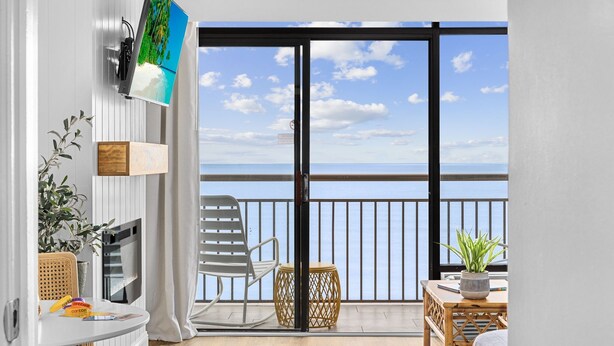 Un balcón con vista al mar, una silla blanca, una mesita redonda y un televisor montado en la pared.