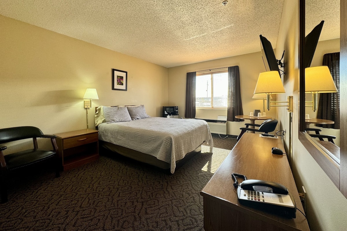 A hotel room with a bed, a desk with a telephone, a chair, a desk lamp, a mirror, and a window with curtains.