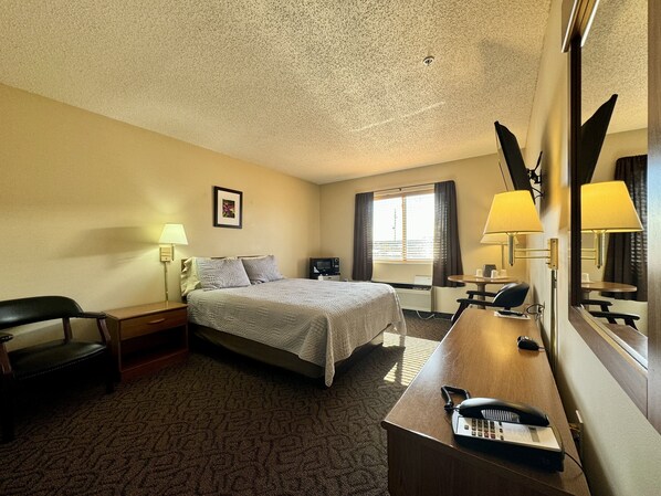 A hotel room with a bed, a desk with a telephone, a chair, a desk lamp, a mirror, and a window with curtains.
