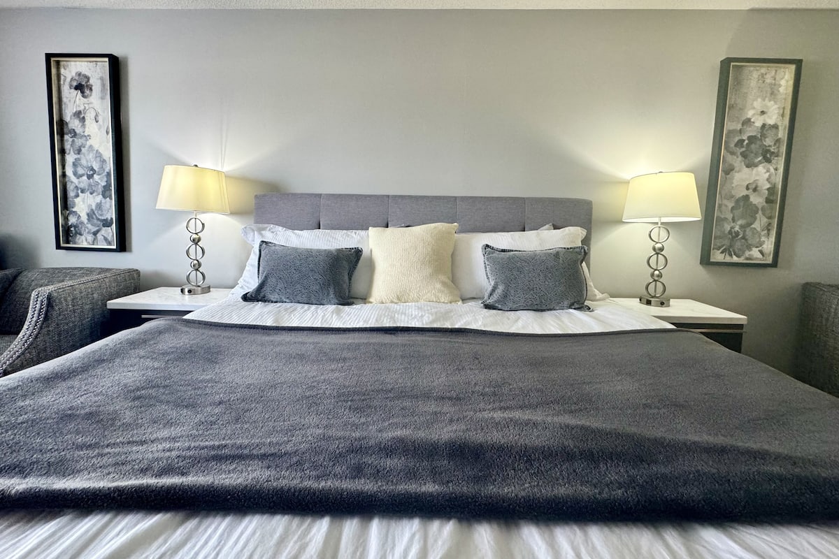 A neatly made bed with a grey headboard, white linens, and grey pillows. There are two bedside tables with lamps and framed floral artwork on the walls.