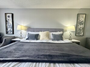 A neatly made bed with a grey headboard, white linens, and grey pillows. There are two bedside tables with lamps and framed floral artwork on the walls.