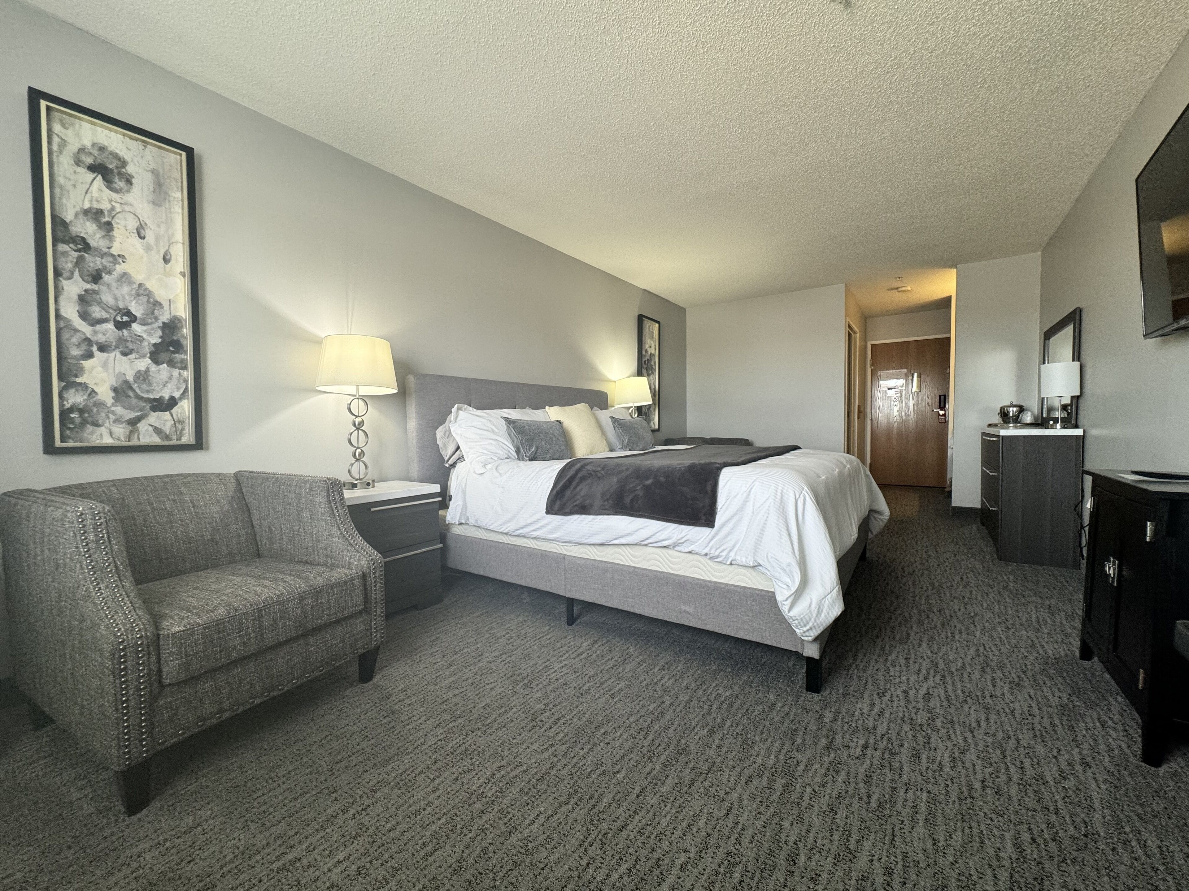A hotel room with a bed, a chair, a nightstand, a dresser, and a painting on the wall.