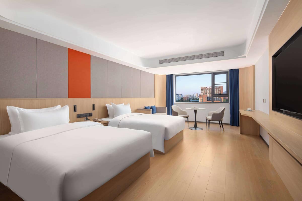 A hotel room with two beds, a desk, a chair, a TV, and a large window with a city view.