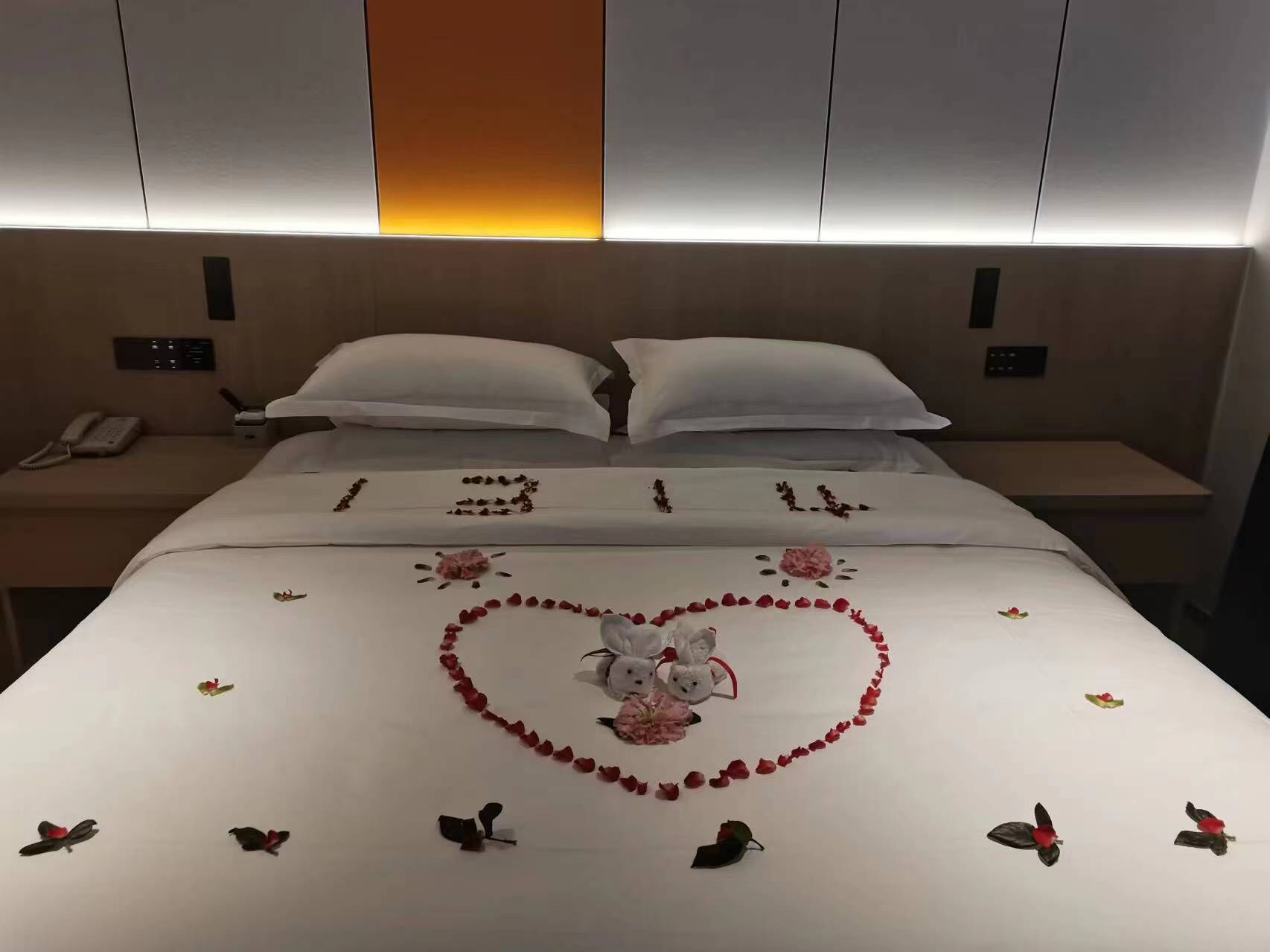 A hotel room with a neatly made bed, a bedside table with a phone, and a wall-mounted control panel.