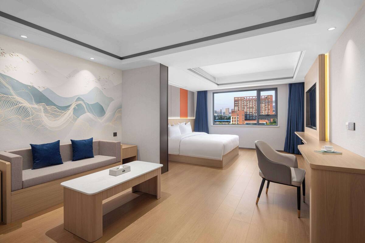 A modern hotel room with a sofa, a bed, a desk, and a view of the cityscape.