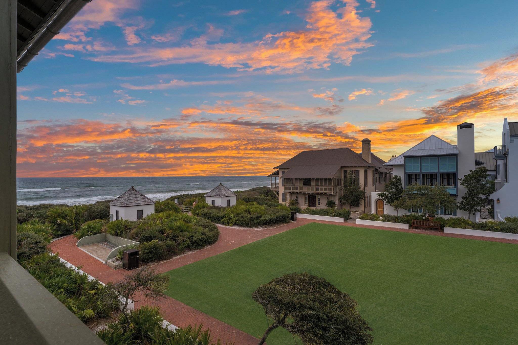 A coastal property with a view of the ocean at sunset, featuring a well-maintained lawn, a small gazebo, and a large building with multiple windows.